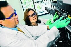 Two people in lab safety gear work on an electronic device near a bright window.