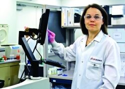 A woman in a lab coat, goggles, and gloves pokes a computer screen in a laboratory.