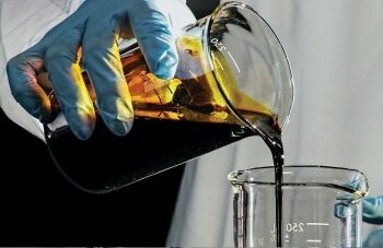 A gloved hand pours a brown-black goo from one beaker into another.